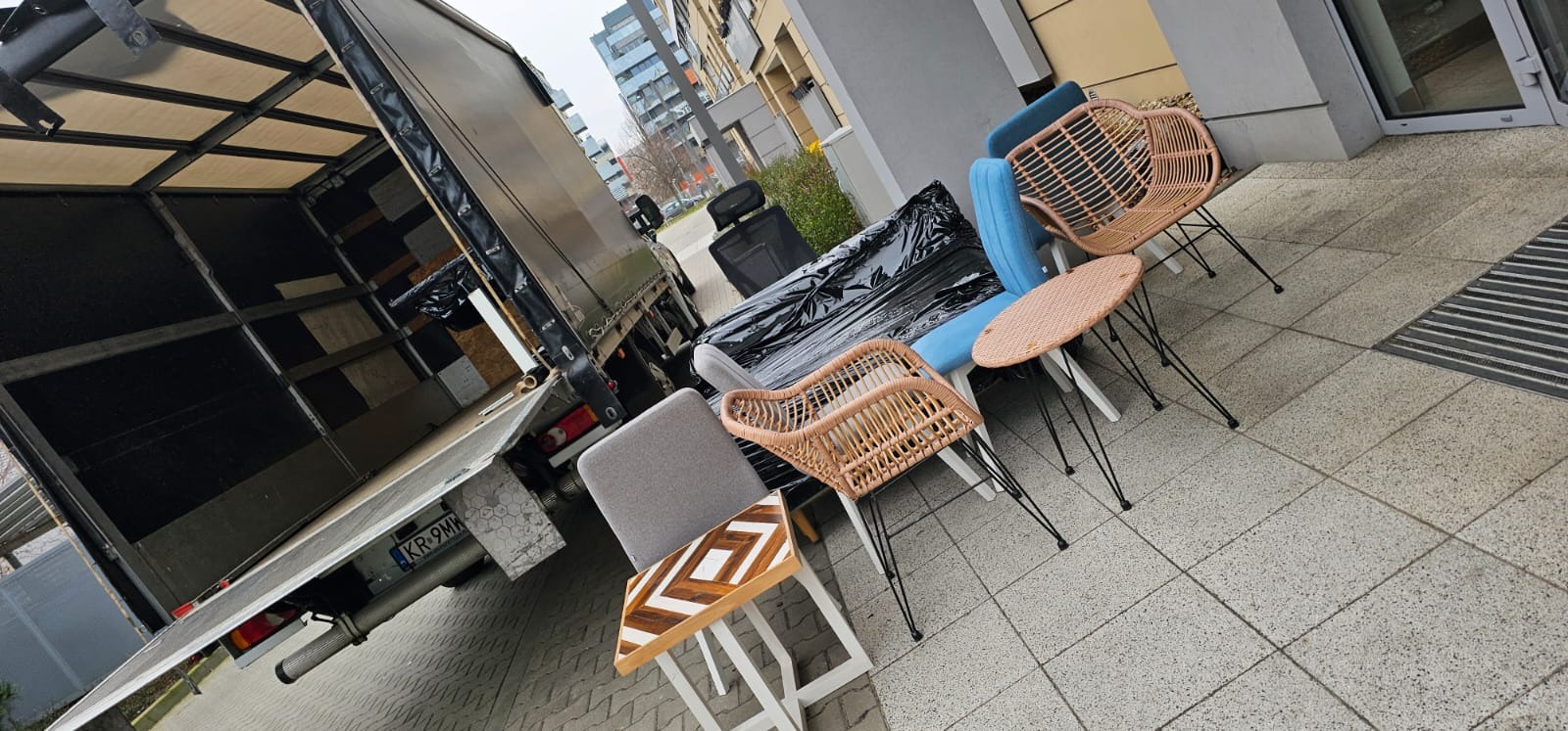 Otwarta paka busa z widocznymi panelami wewnętrznymi, obok wystawione meble ogrodowe i biurowe przygotowane do załadunku na chodniku przed budynkiem mieszkalnym.