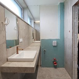 Iwona Pabian - Wnętrze łazienki w trakcie prac wykończeniowych: zamontowana umywalka z baterią, lustro, częściowo położone płytki, widoczne instalacje.