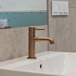 Iwona Pabian - Bateria umywalkowa w odcieniu miedzi na tle białej umywalki i płytek imitujących marmur. Widoczne elementy instalacji i lustro z niedokończonym wykończeniem.