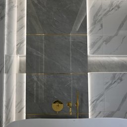 Iwona Pabian - Nowoczesna łazienka z wanną wolnostojącą, szarymi i białymi płytkami imitującymi marmur, złotą armaturą i subtelnym oświetleniem LED.