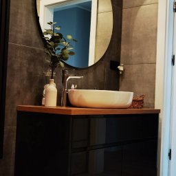Iwona Pabian - Nowoczesna łazienka z okrągłym lustrem, umywalką nablatową i czarną szafką. Szare płytki na ścianach i podłodze. Minimalistyczny design z roślinnym akcentem.