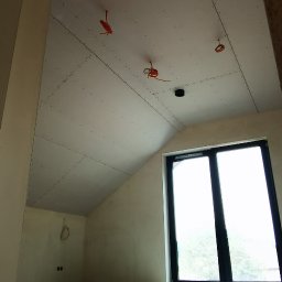 Iwona Pabian - Poddasze z oknem, ściany i skośny sufit pokryte płytami kartonowo-gipsowymi, widoczne kable elektryczne. Etap prac wykończeniowych.