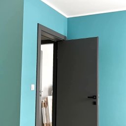 Iwona Pabian - Turkusowe ściany i ciemne drzwi w nowym domu. Widoczne panele podłogowe i biały sufit. Minimalistyczny design, świeżo po malowaniu, w trakcie wykańczania.