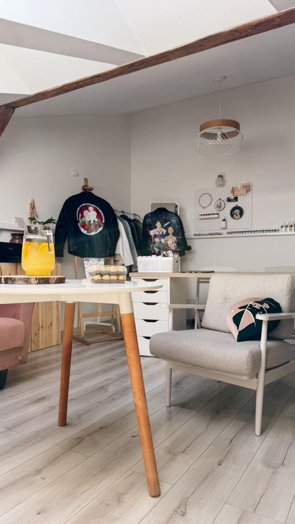 Jasne wnętrze pokoju z drewnianą podłogą, stołem z napojem i przekąskami, krzesłem oraz dekoracyjnymi kurtkami na wieszakach. Widoczna lampa sufitowa.