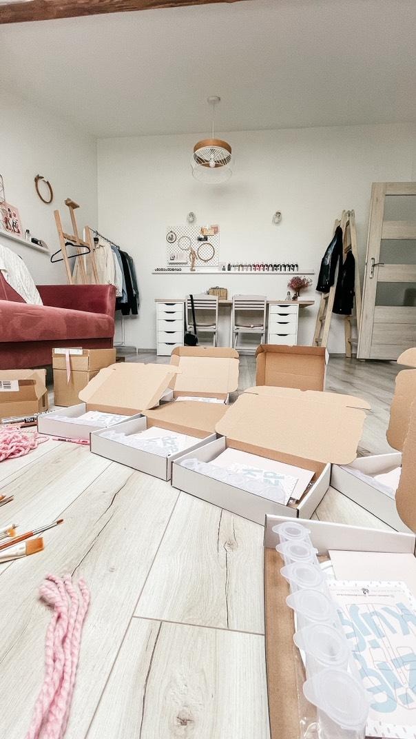 Jasne wnętrze z drewnianą podłogą, gdzie rozłożone są otwarte kartony z zestawami do rękodzieła, pędzle i włóczka. W tle biurko i sofa.