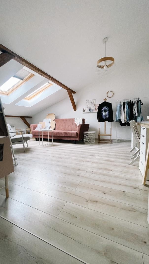 Jasne panele podłogowe w pokoju na poddaszu z oknami dachowymi i różową sofą. Widoczny fragment garderoby i lampa sufitowa.