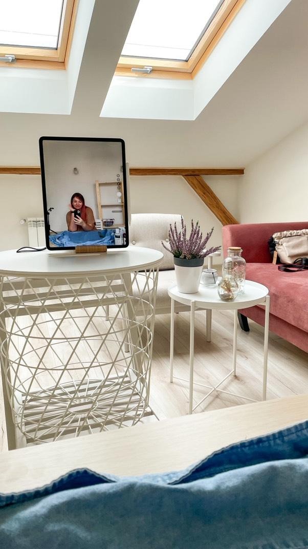 Jasne poddasze z oknami dachowymi, drewnianą belką i różową sofą. Na pierwszym planie stoliki z dekoracjami i odbicie kobiety w lustrze.