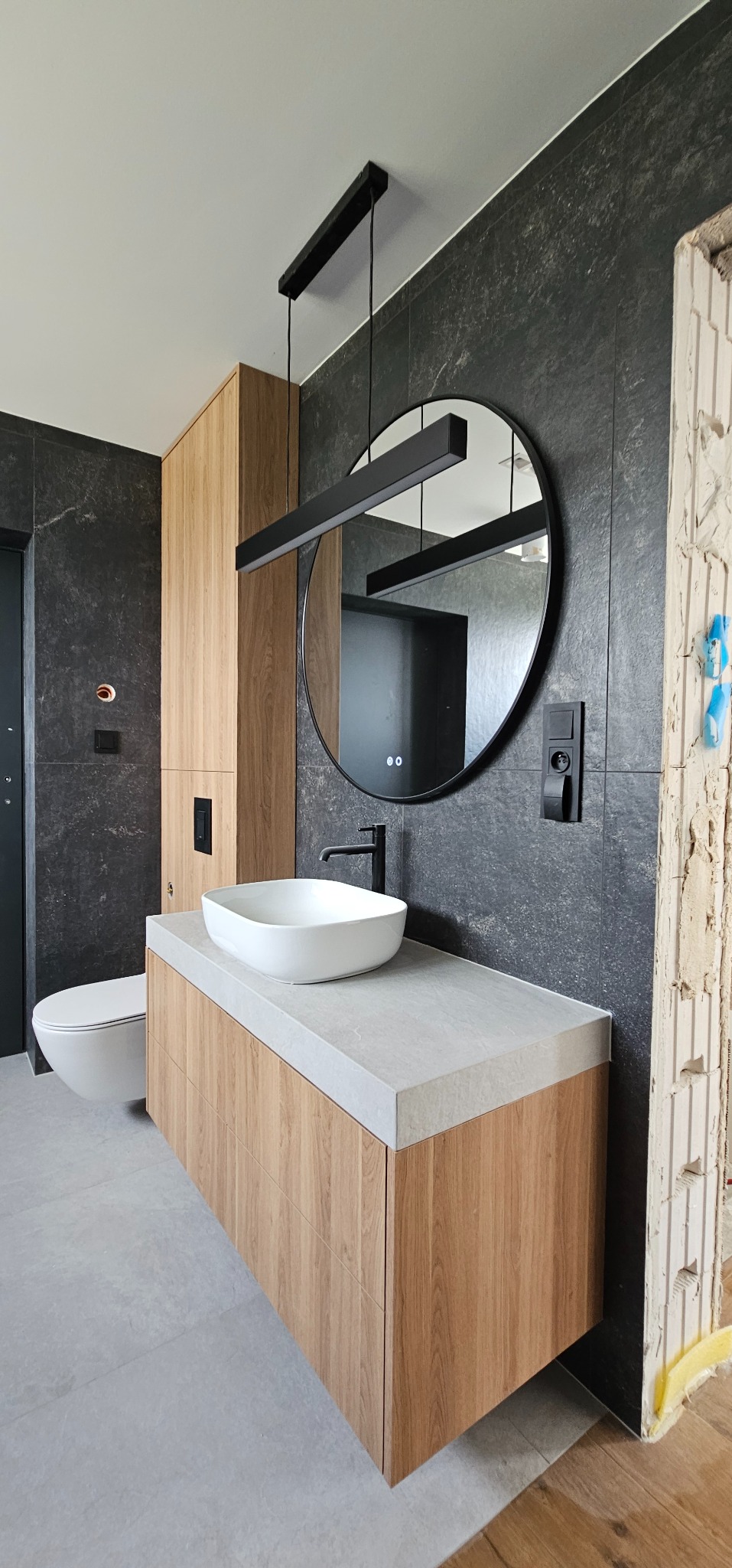 Nowoczesna łazienka z czarnymi płytkami, drewnianą szafką pod umywalką i okrągłym lustrem z podświetleniem LED. Fragment ściany w stanie surowym.