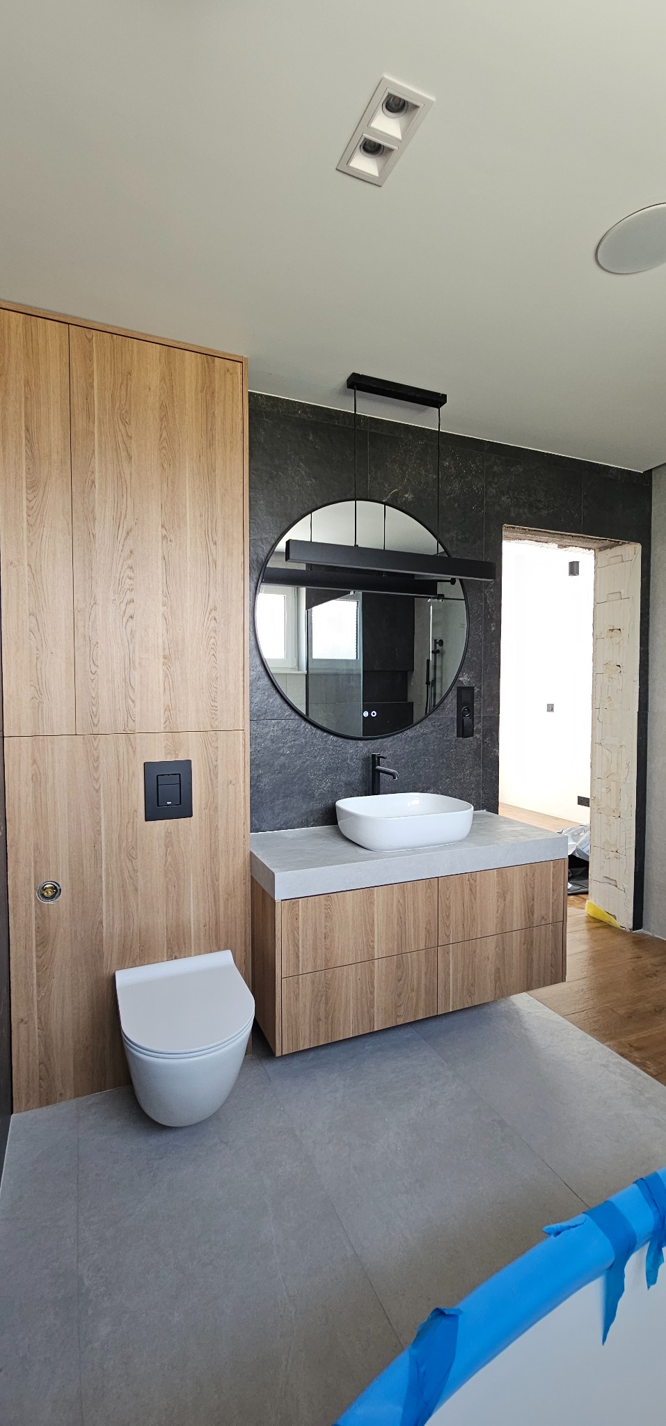 Nowoczesna łazienka w Gliwicach: drewniane szafki, okrągłe lustro z podświetleniem, szare płytki i wisząca toaleta. Minimalistyczny design i eleganckie wykończenie.