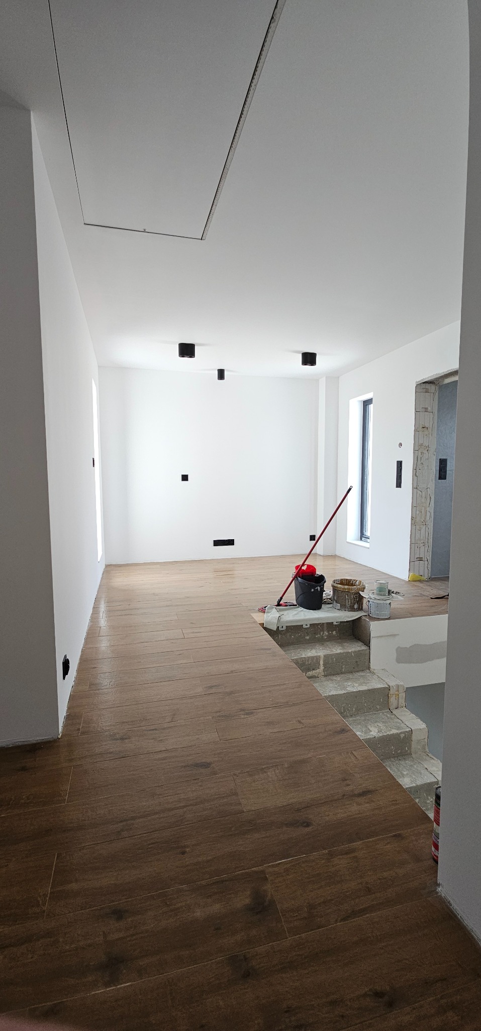 Jasne wnętrze z drewnopodobną podłogą, widoczne schody w stanie surowym i akcesoria malarskie. Minimalistyczne oświetlenie sufitowe.