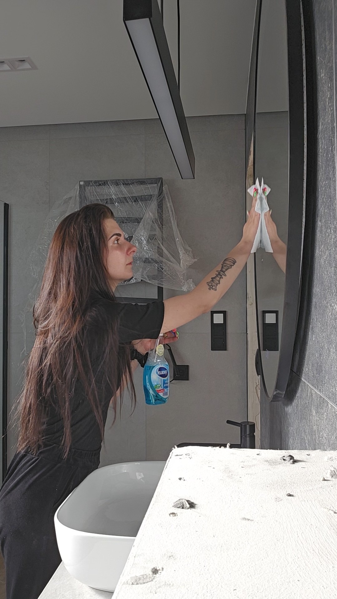 Kobieta czyści okrągłe lustro w łazience po remoncie, używając płynu i ściereczki. Widoczna umywalka, szare płytki i czarne elementy armatury.