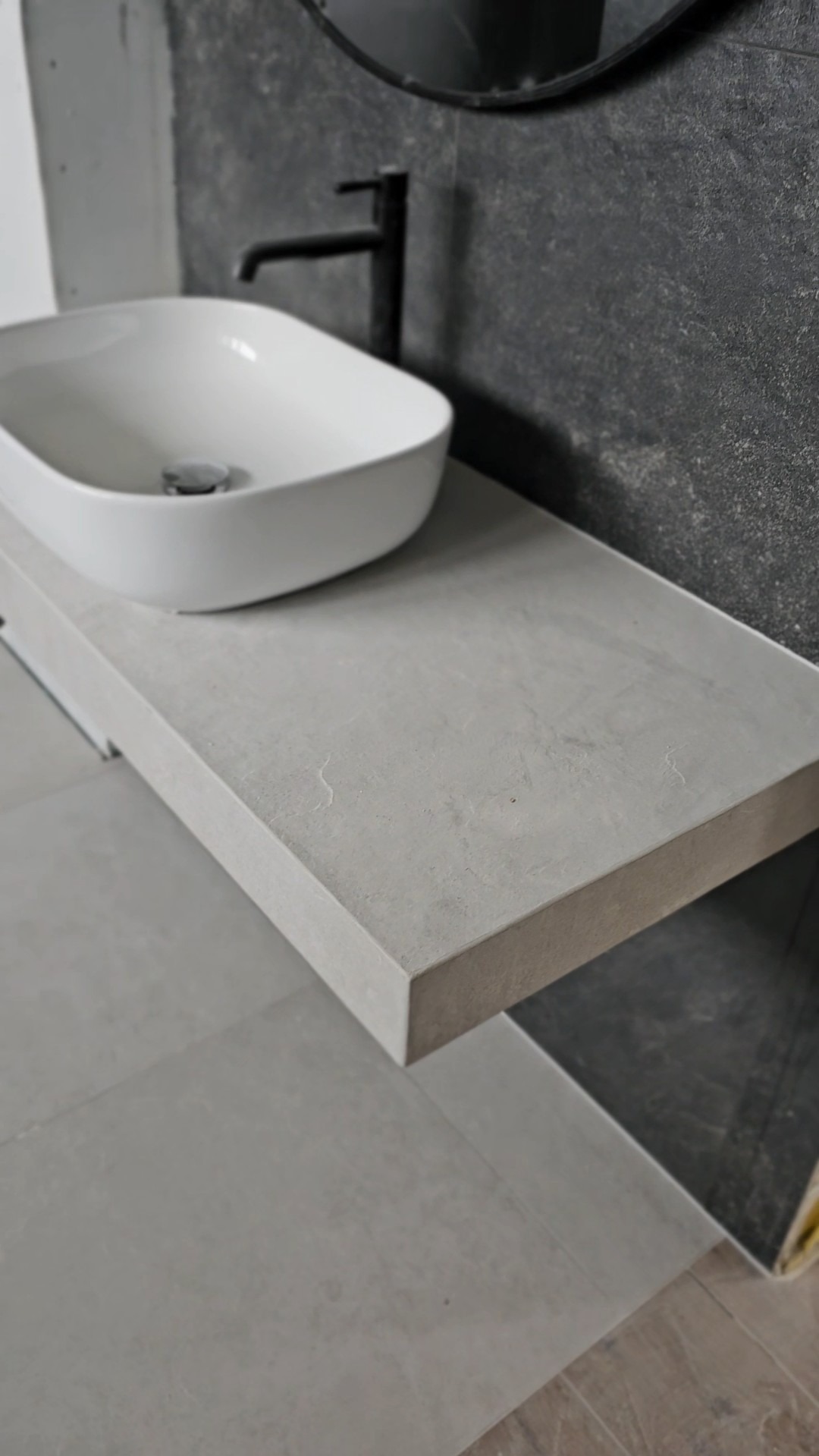 Nowoczesna łazienka: biała umywalka nablatowa na szarym blacie z konglomeratu, czarna bateria, szara ściana z efektem kamienia i okrągłe lustro w tle.