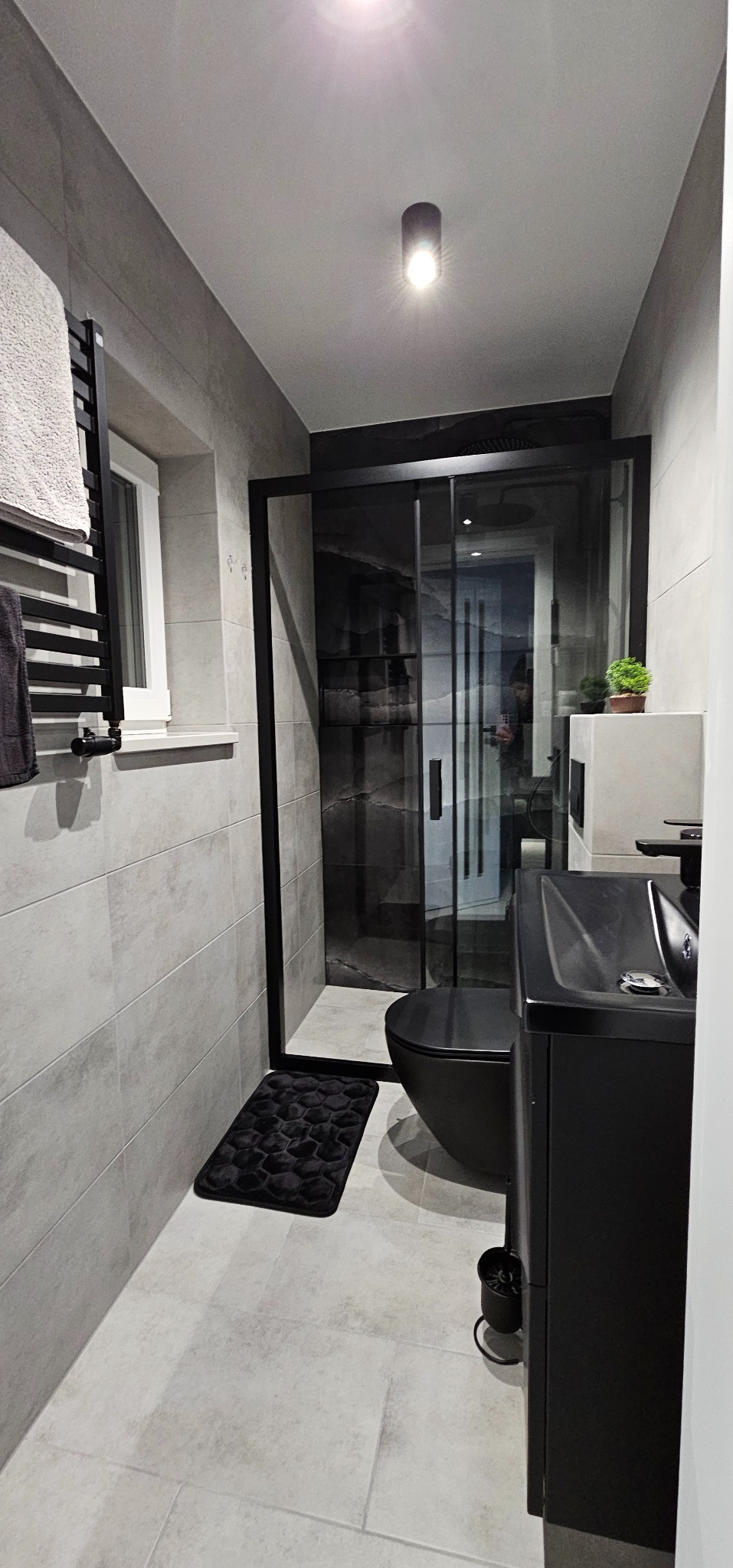 Nowoczesna łazienka w szarościach z czarnymi akcentami: kabina prysznicowa, umywalka z szafką, toaleta. Minimalistyczny design, płytki na ścianach i podłodze.