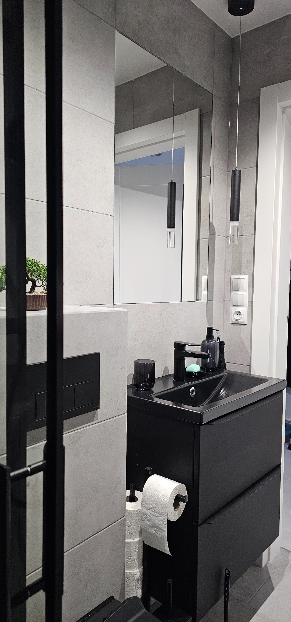Nowoczesna łazienka z czarną szafką umywalkową, szarą glazurą i minimalistycznym oświetleniem. W lustrze odbija się fragment białych drzwi.