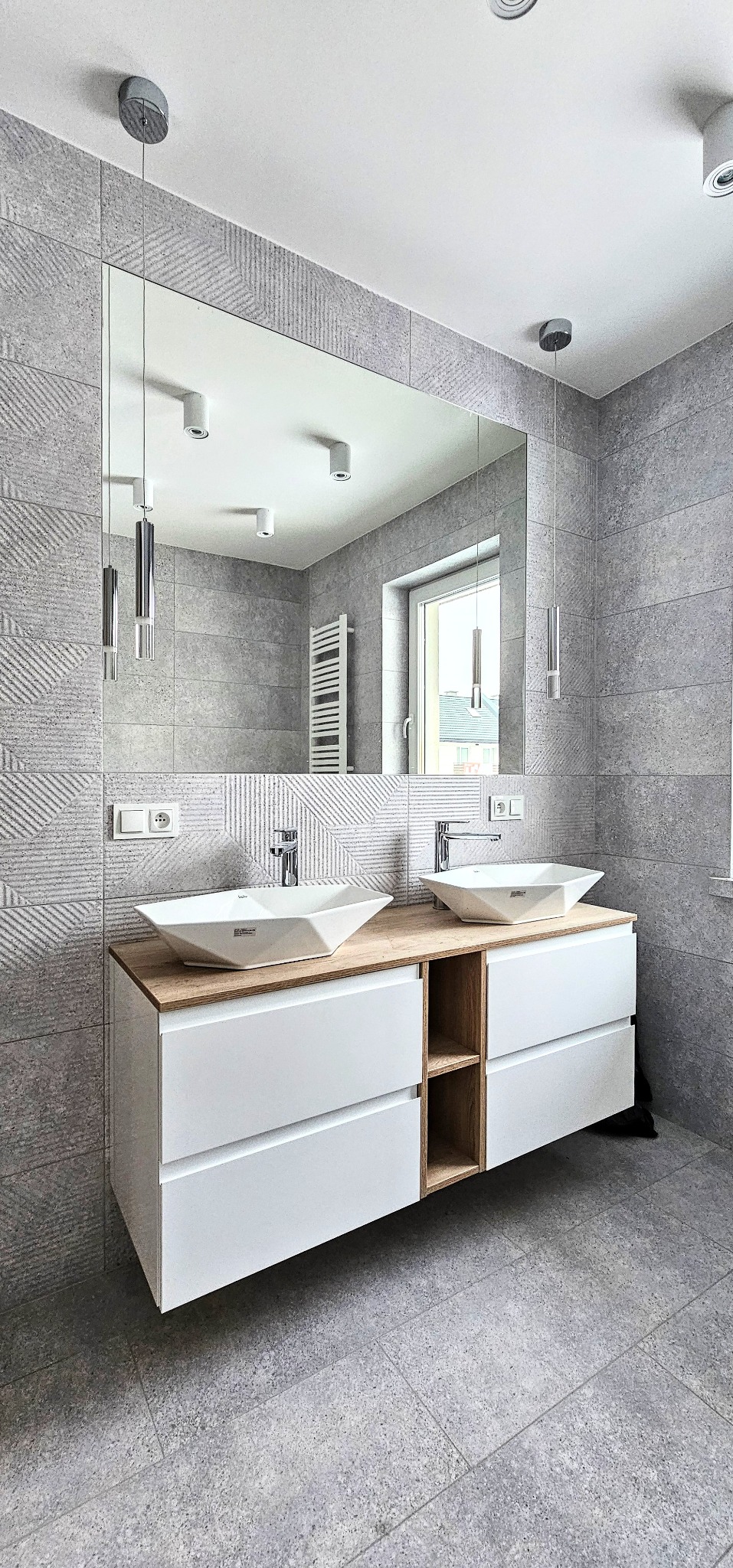 Nowoczesna łazienka w szarościach: podwójna umywalka nablatowa, duże lustro, dekoracyjne płytki ścienne i minimalistyczne oświetlenie.