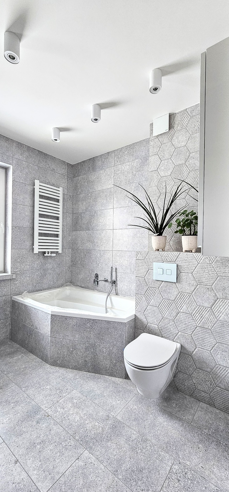 Szara łazienka z wanną narożną, toaletą podwieszaną i grzejnikiem drabinkowym. Heksagonalne płytki na ścianie, rośliny w doniczkach. Nowoczesne oświetlenie sufitowe.