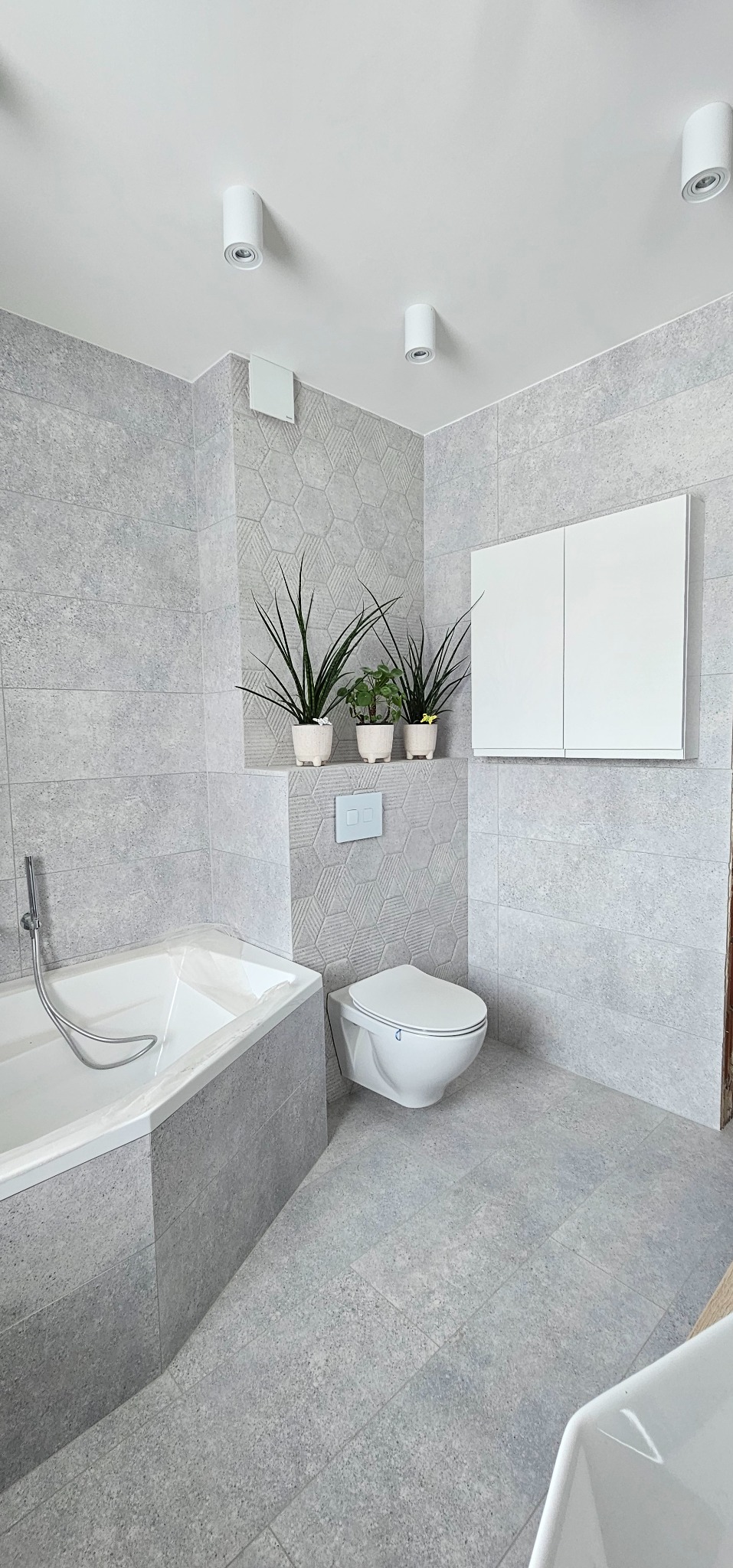 Jasna łazienka po remoncie z szarymi płytkami, wanną, podwieszaną toaletą i roślinami doniczkowymi. Minimalistyczny design z akcentem natury.