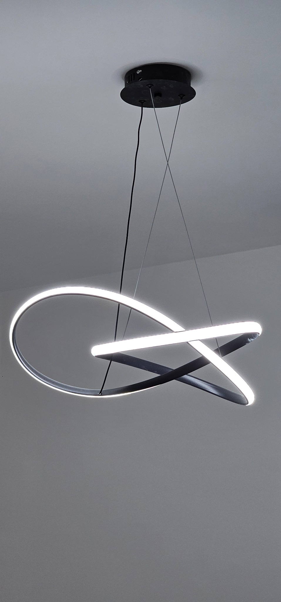 Nowoczesna lampa sufitowa LED z czarną obudową i białym, geometrycznym światłem, zawieszona na tle szarego sufitu. Minimalistyczny design oświetlenia.