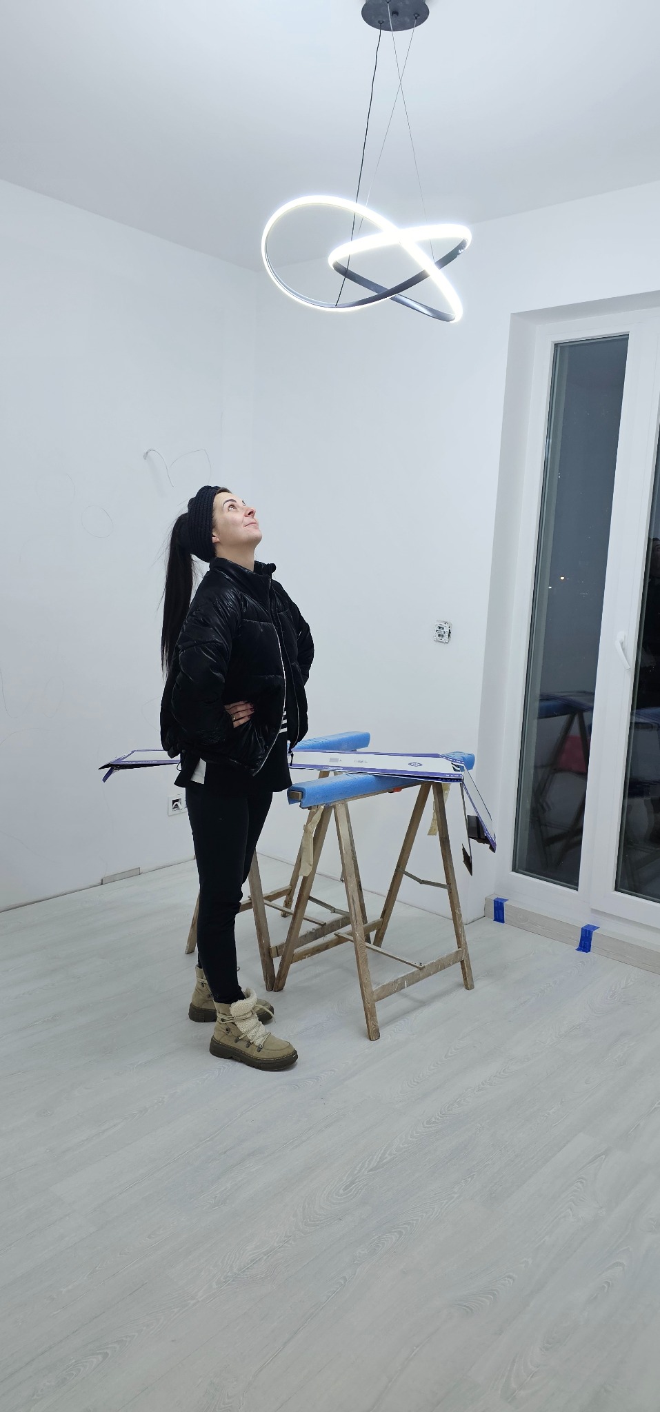 Kobieta spogląda na nowoczesną lampę sufitową w kształcie abstrakcyjnej spirali w jasnym, odnowionym wnętrzu. Widoczne narzędzia malarskie i okno.