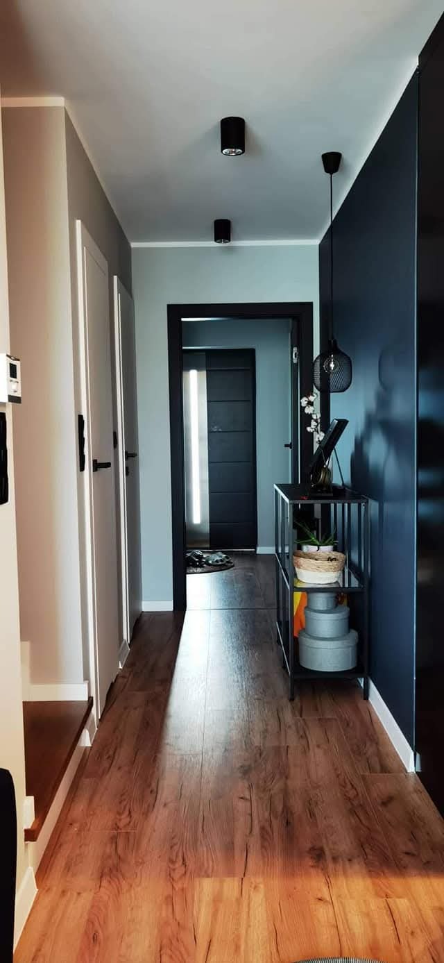 Nowoczesny korytarz w domu deweloperskim: drewnopodobne panele, szara ściana z białymi drzwiami, granatowa ściana z czarną konsolą i lampą wiszącą.