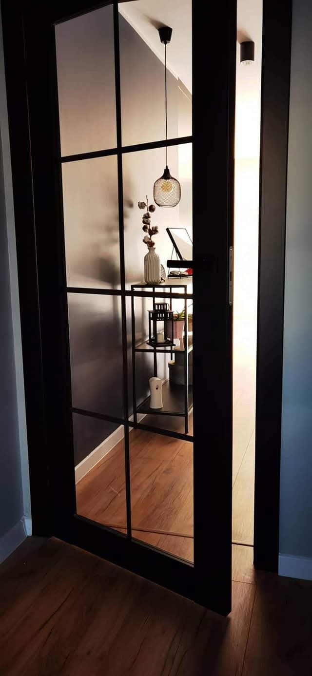 Nowoczesne drzwi przesuwne z czarną ramą i szklanymi panelami, widok na wnętrze z drewnianą podłogą, metalowym regałem i dekoracyjnym oświetleniem.