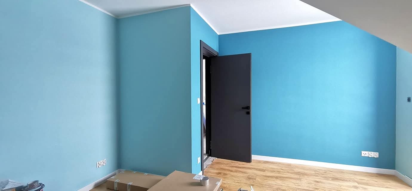 Błękitne ściany w nowym domu deweloperskim, drewnopodobna podłoga, czarne drzwi wewnętrzne, białe listwy przypodłogowe i sufitowe. Widoczne gniazdka elektryczne.