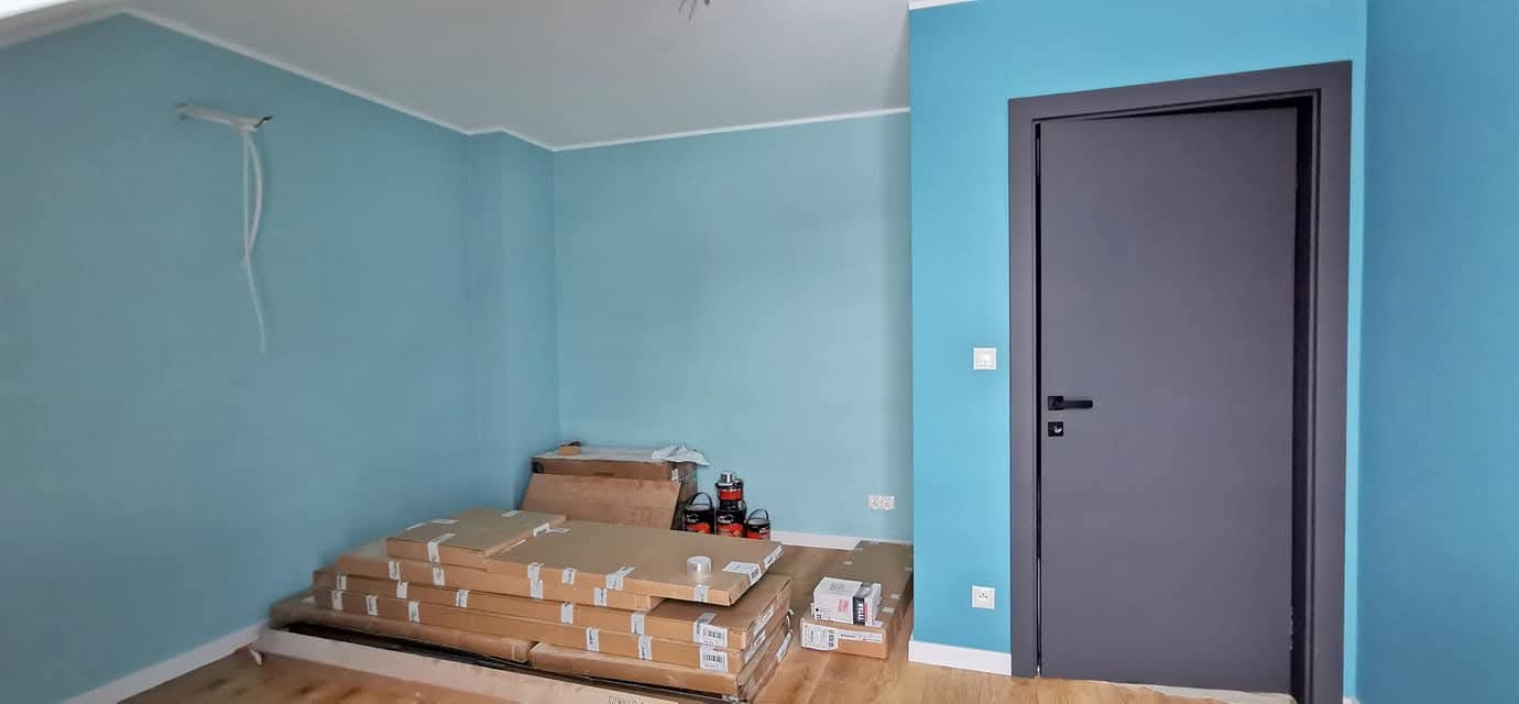 Niebieskie ściany w pokoju z szarą ościeżnicą i drzwiami. Na podłodze leżą kartony z panelami. Widoczne puszki z farbą. Dom w stanie deweloperskim.