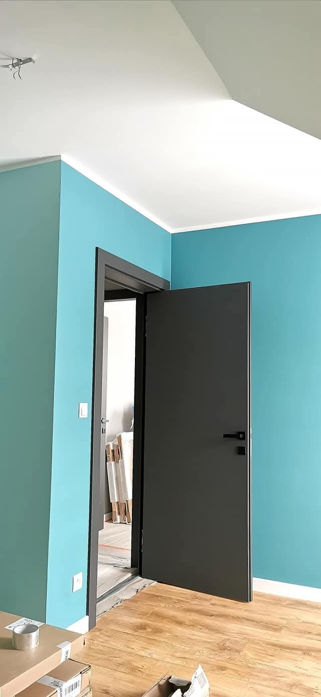 Turkusowe ściany i ciemne drzwi w nowym domu. Widoczne panele podłogowe i biały sufit. Minimalistyczny design, świeżo po malowaniu, w trakcie wykańczania.