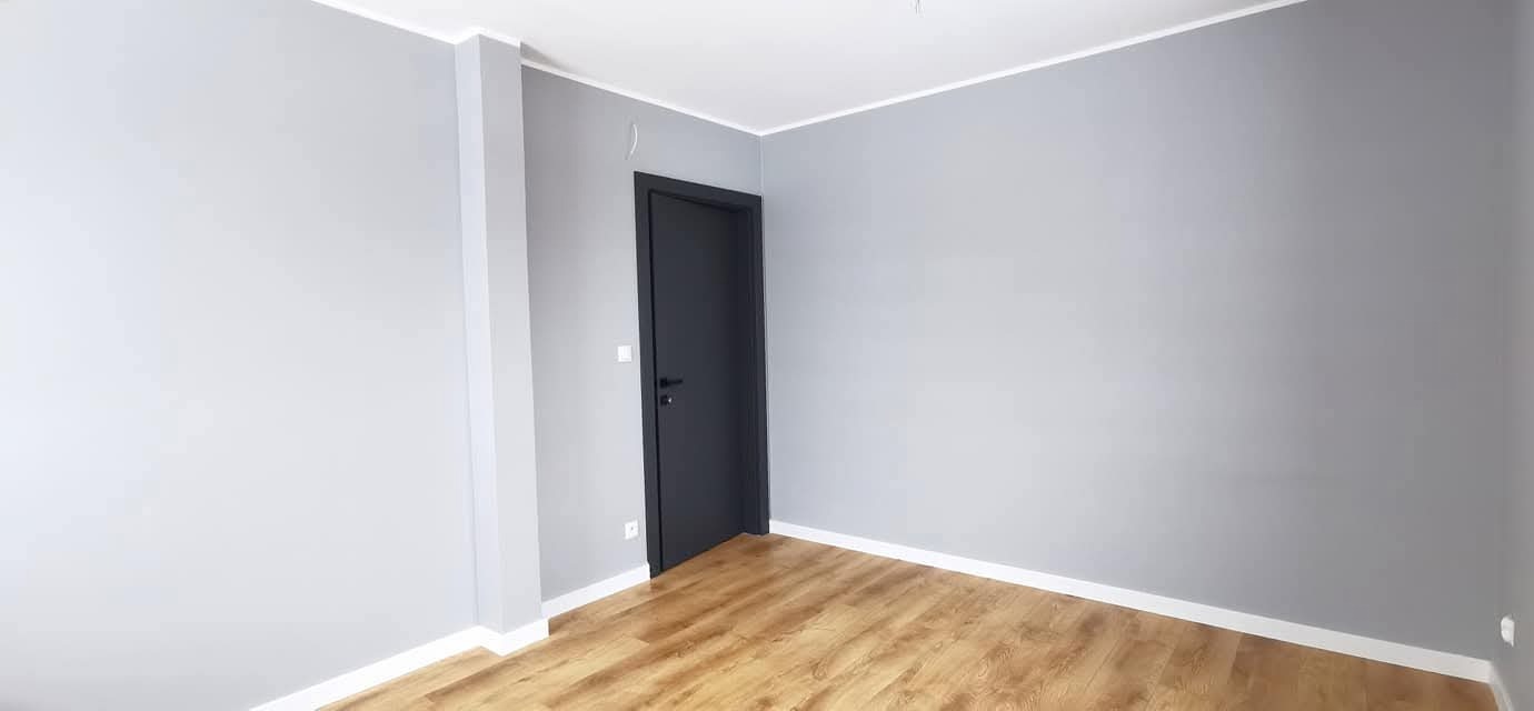 Szare ściany, drewnopodobne panele i ciemne drzwi w nowym, pustym pokoju. Białe listwy przypodłogowe i sufitowe. Minimalistyczne wykończenie wnętrza.