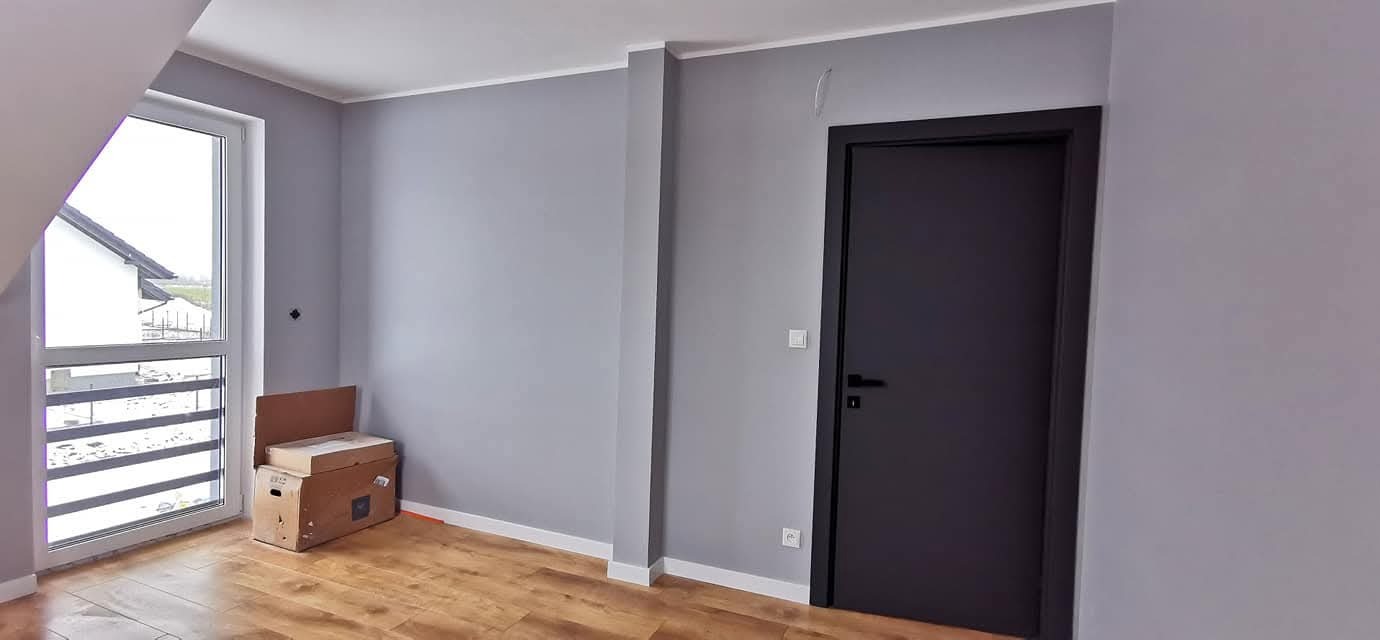Szare ściany w nowym domu deweloperskim, drewnopodobne panele, czarne drzwi i białe listwy przypodłogowe. Kartony przy oknie. Minimalistyczne wykończenie.