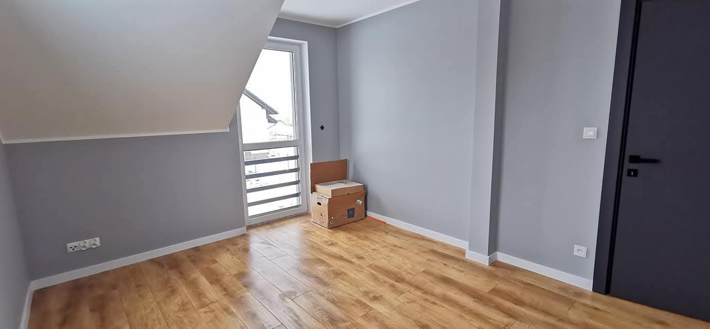 Jasne panele podłogowe w pokoju z szarymi ścianami i oknem. Widoczne kartony, listwy przypodłogowe i ciemne drzwi. Pokój w domu deweloperskim po wykończeniu.