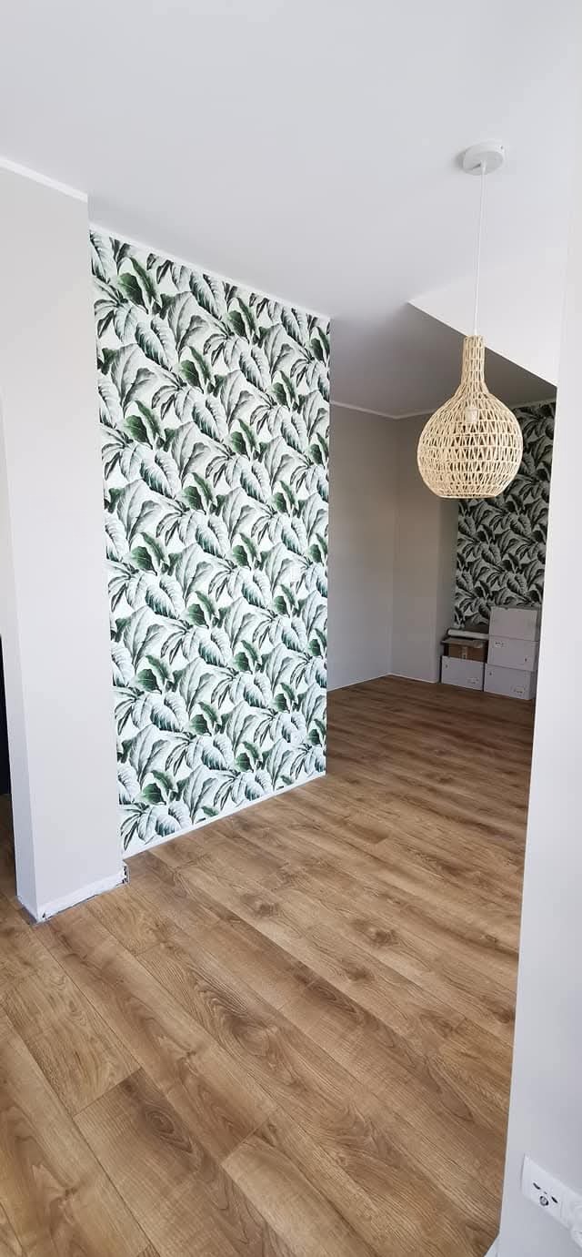 Jasne panele podłogowe i ściana z tapetą w liście w nowym domu w Gliwicach. W tle widoczna lampa z abażurem z wikliny i kartony.