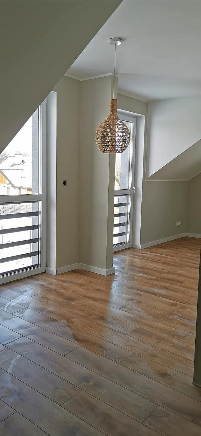 Jasne wnętrze poddasza z drewnianą podłogą i oknami balkonowymi. W centrum wisi lampa z wikliny. Ściany w odcieniach beżu, widoczne skosy.