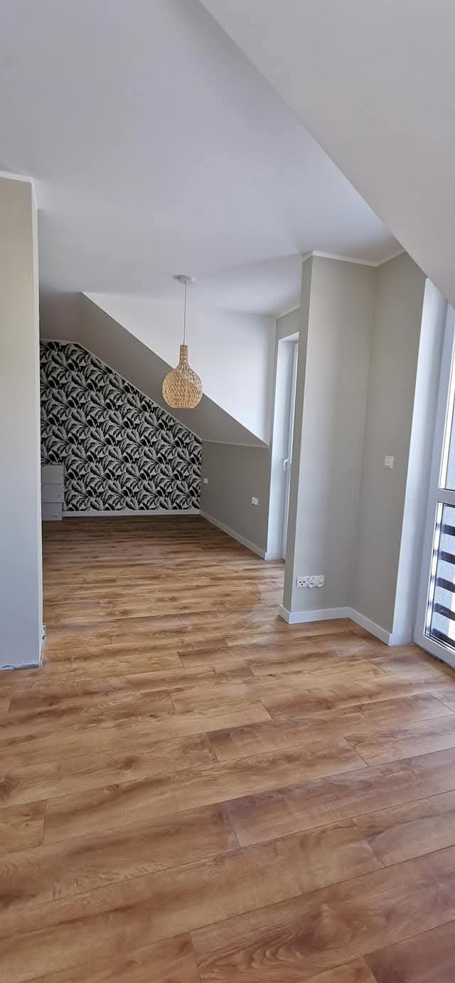 Jasne panele podłogowe w pokoju na poddaszu z wzorzystą tapetą na skośnej ścianie. W tle widoczne białe szafki i okno balkonowe.