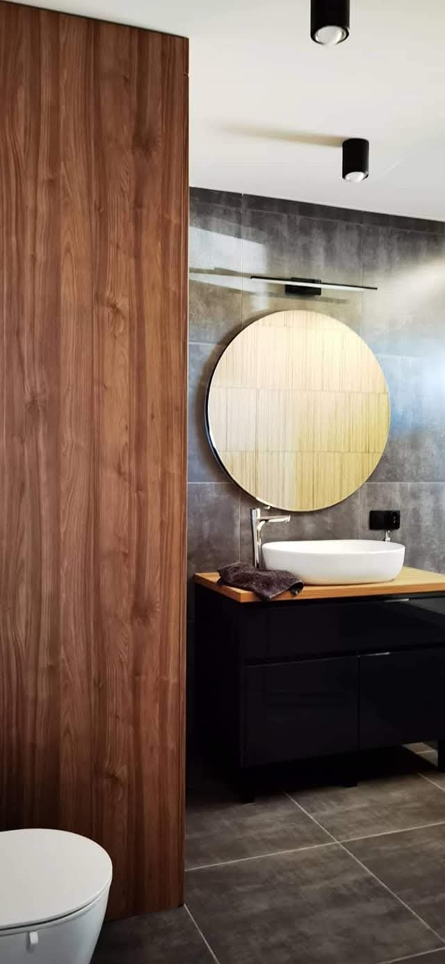 Nowoczesna łazienka z drewnianą ścianą, okrągłym lustrem i czarną szafką pod umywalką. Szare płytki na ścianach i podłodze. Minimalistyczne oświetlenie sufitowe.