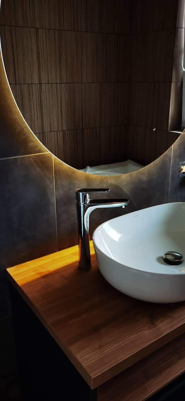 Nowoczesna łazienka: biała umywalka nablatowa z chromowaną baterią na drewnianym blacie, okrągłe lustro z podświetleniem i ciemne płytki imitujące drewno.