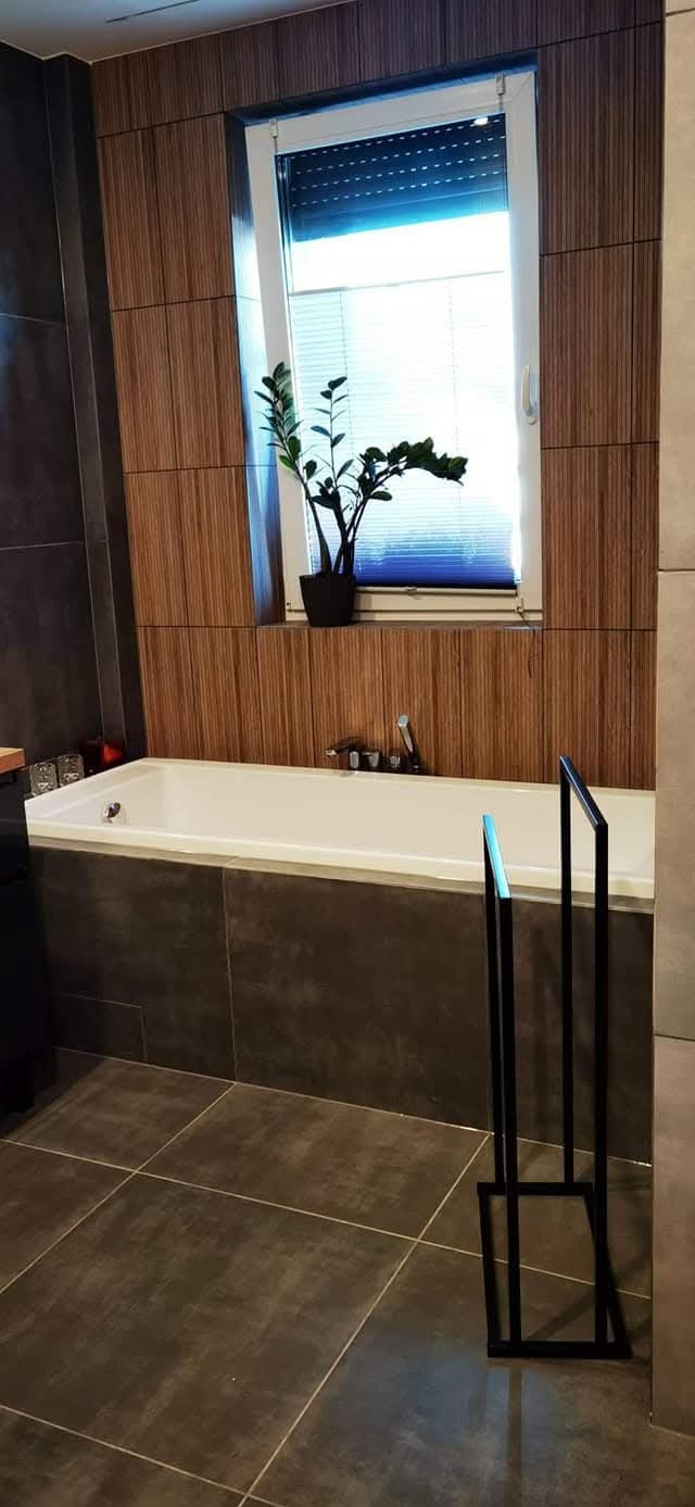 Nowoczesna łazienka z wanną obudowaną ciemnymi płytkami, drewnopodobne panele na ścianie, roślina w oknie i czarny wieszak na ręczniki.
