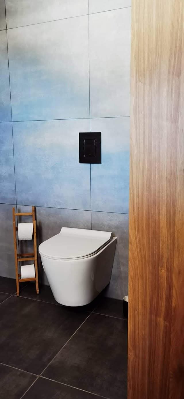 Nowoczesna łazienka z podwieszaną toaletą, błękitnymi płytkami i drewnianym panelem. Minimalistyczny design, ciemna podłoga i drewniany regał na papier toaletowy.