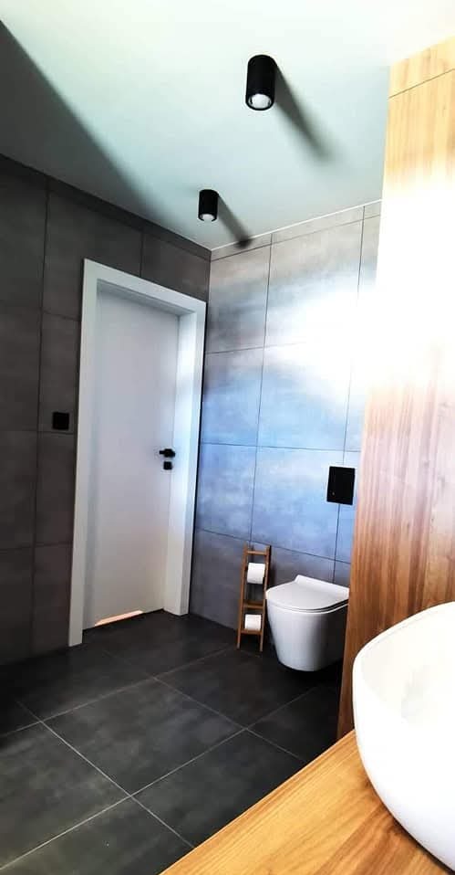 Nowoczesna łazienka w szarościach z drewnianymi akcentami. Uwagę przykuwają duże, kwadratowe płytki na ścianach i podłodze oraz minimalistyczna armatura. Widoczna toaleta i umywalka.