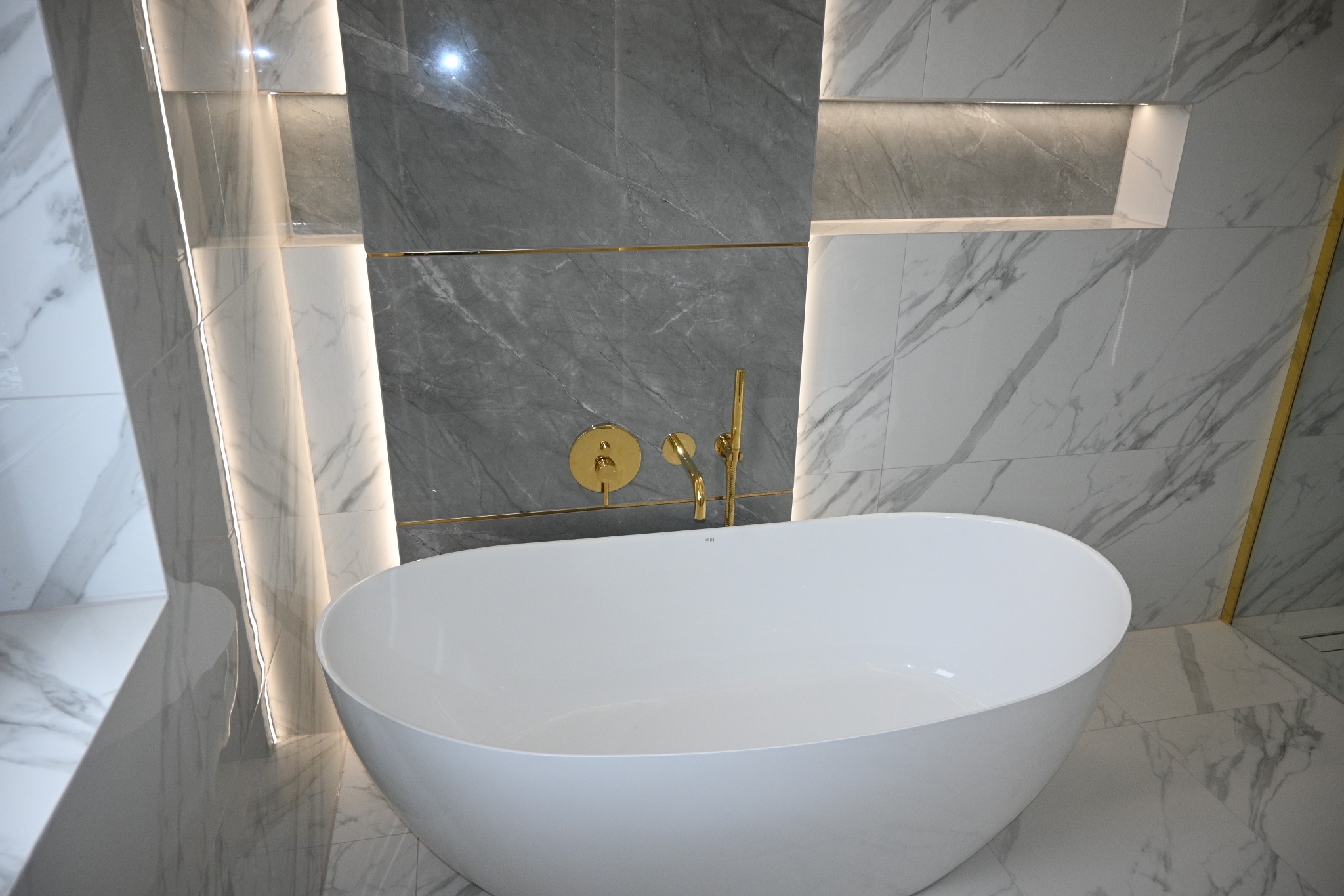 Nowoczesna łazienka z wolnostojącą białą wanną, złotą armaturą i marmurowymi płytkami w odcieniach szarości. Podświetlane wnęki dodają elegancji.