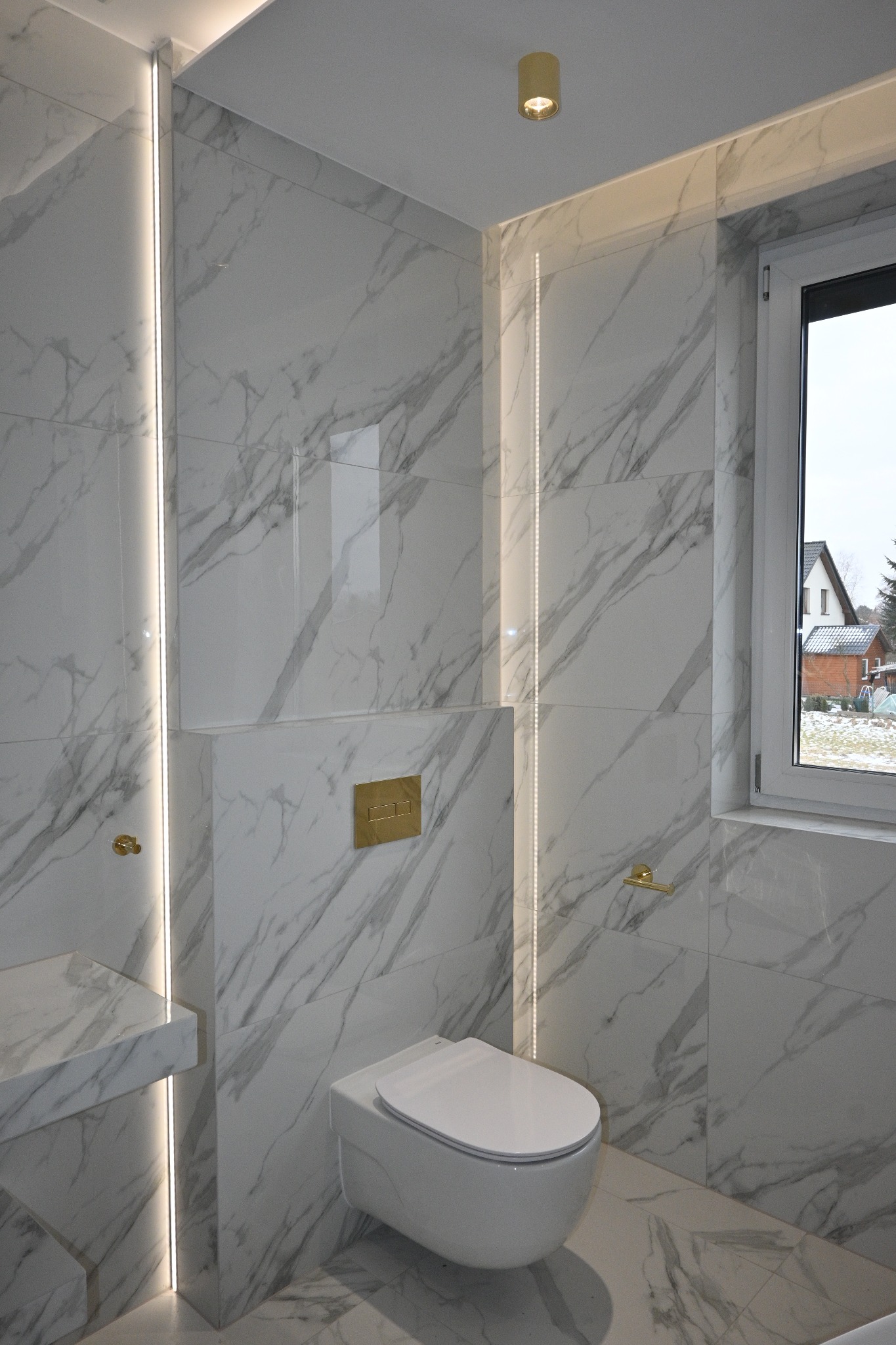 Elegancka łazienka z białymi płytkami imitującymi marmur, podświetleniem LED i złotymi akcentami. Wisząca toaleta i okno z widokiem na dom.