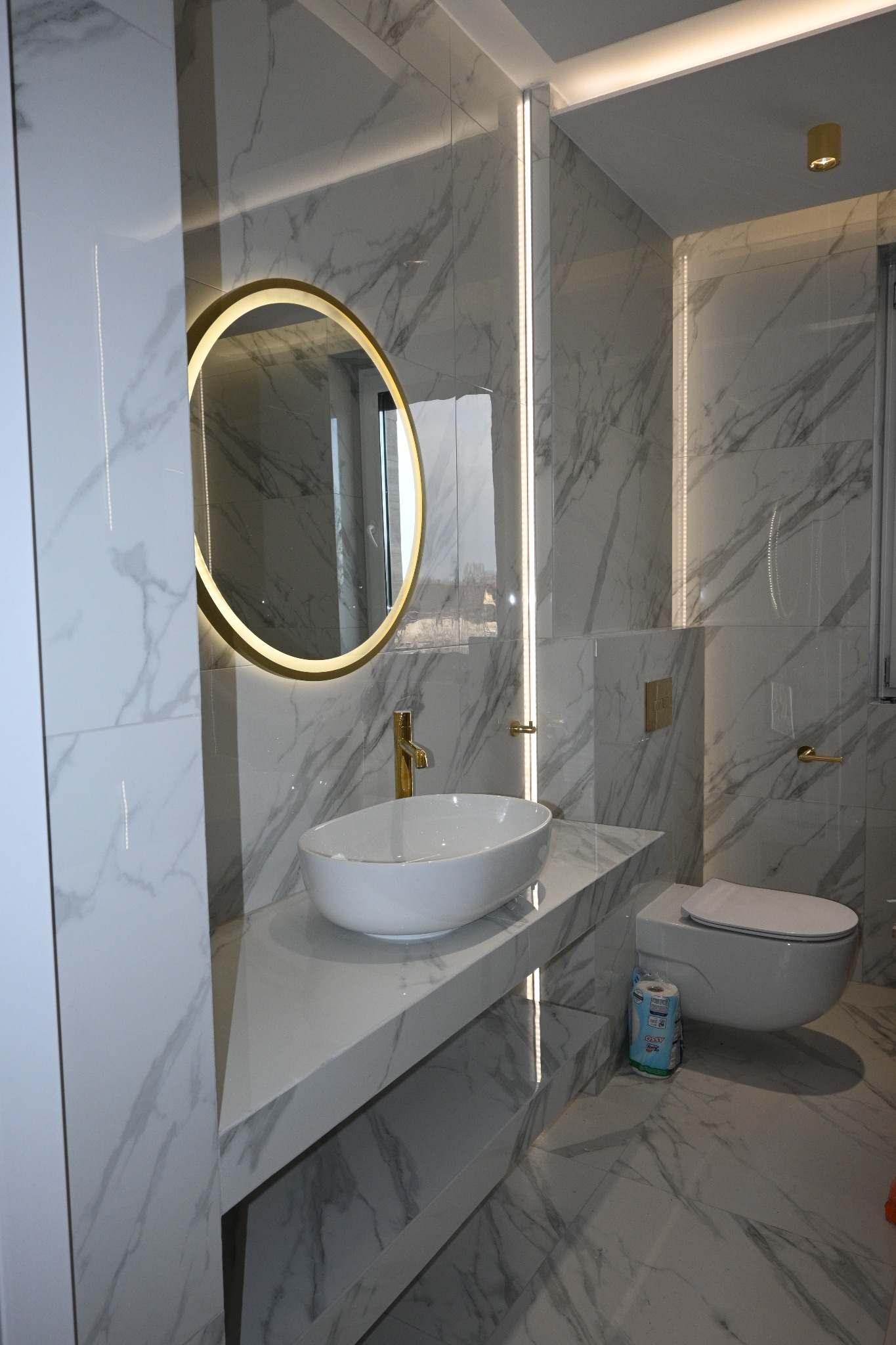 Elegancka łazienka z marmurowymi płytkami, okrągłym lustrem z podświetleniem LED, białą umywalką nablatową i złotą armaturą. Nowoczesne oświetlenie liniowe.