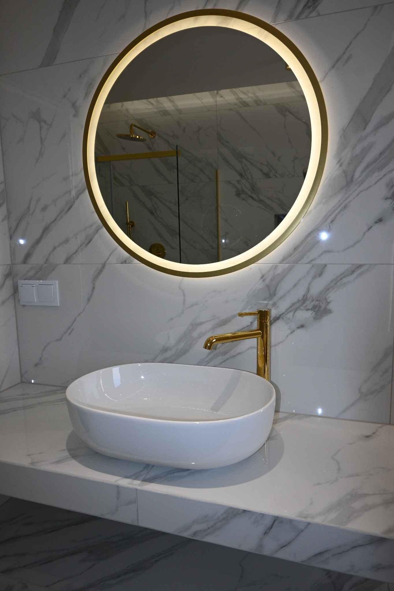 Elegancka umywalka nablatowa z baterią w kolorze złota i okrągłym lustrem z podświetleniem LED na tle marmurowych płytek w łazience w Gliwicach.