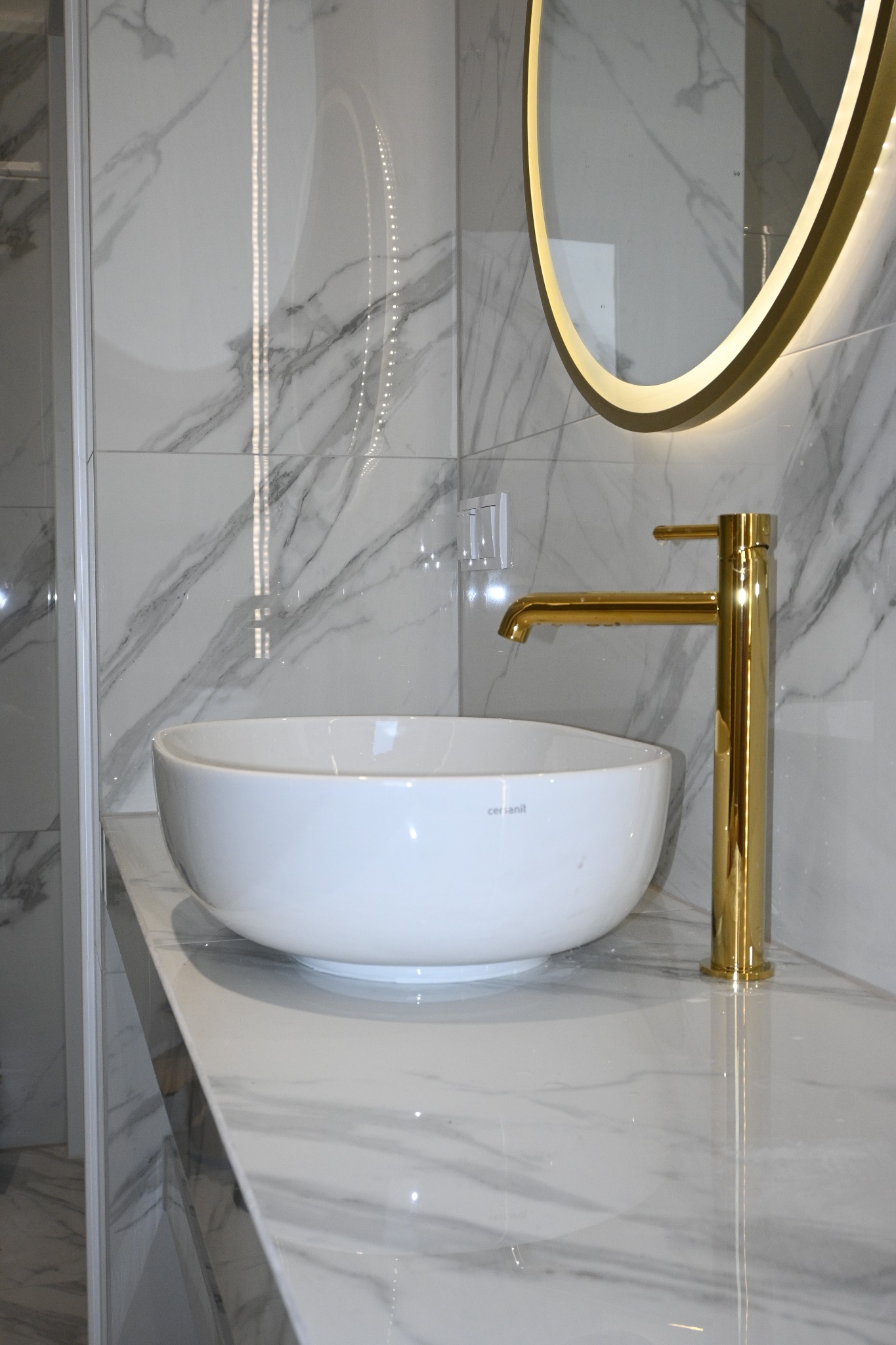 Biała, okrągła umywalka Cersanit z złotą baterią na blacie z marmuru. W tle lustro z podświetleniem LED i ściana z płytek imitujących marmur.