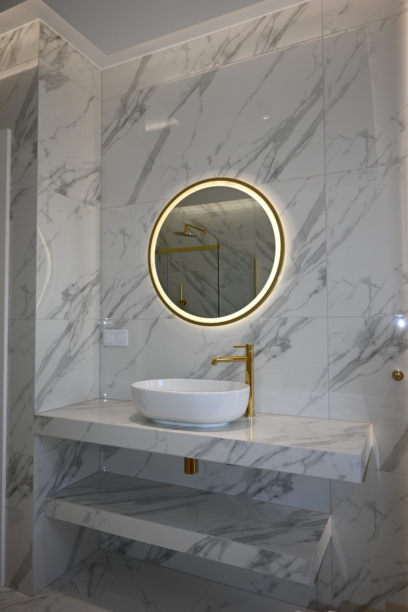 Nowoczesna łazienka: białe marmurowe płytki, okrągłe lustro z podświetleniem LED, biała umywalka nablatowa i złota armatura. Minimalistyczny design.