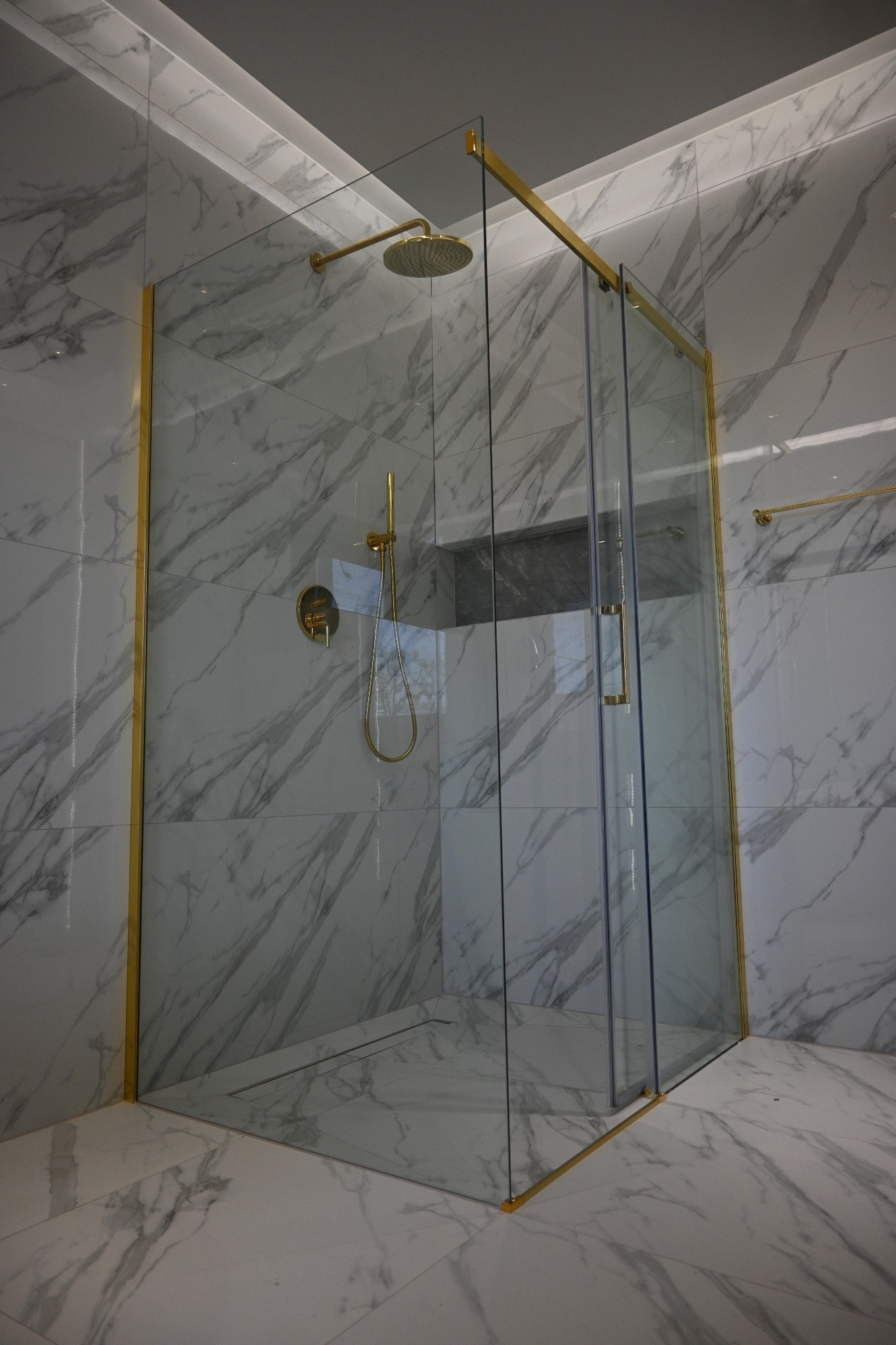 Nowoczesna kabina prysznicowa ze złotymi detalami i marmurowymi płytkami w wykończonej łazience. Widoczny odpływ liniowy i deszczownica.