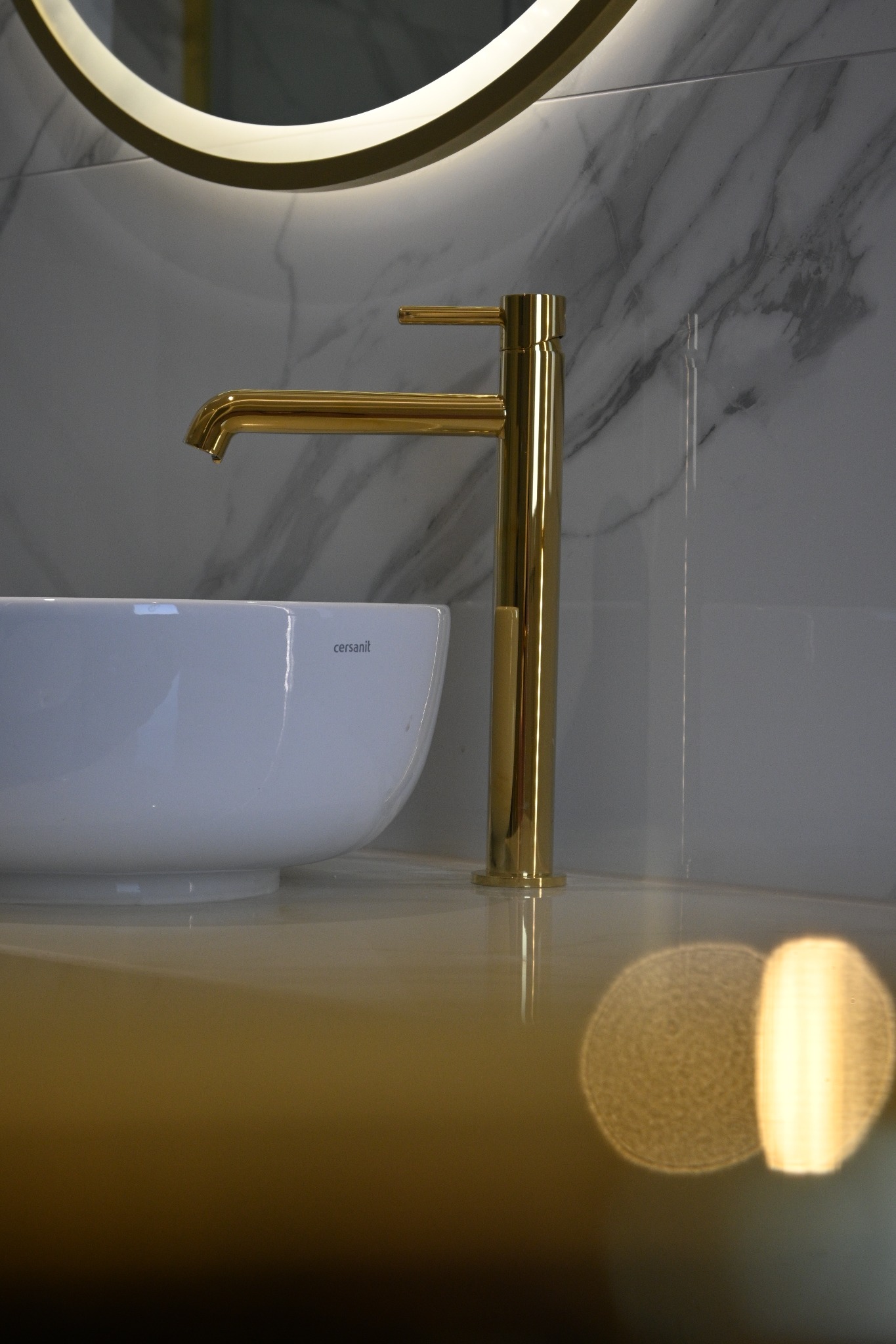 Biała umywalka Cersanit z złotą baterią na blacie, podświetlane okrągłe lustro i marmurowe płytki w tle. Nowoczesna aranżacja łazienki.