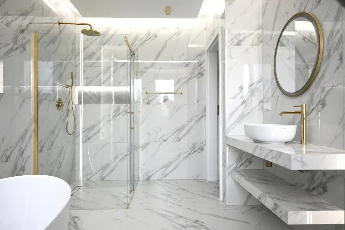 Elegancka łazienka z białymi marmurowymi płytkami, złotymi akcentami armatury i kabiny prysznicowej, okrągłe lustro nad umywalką, nowoczesny design.