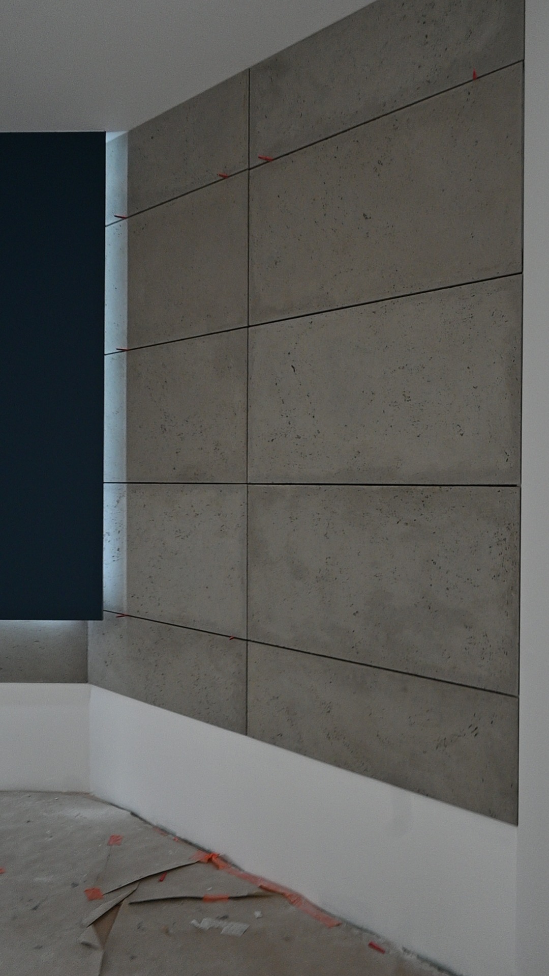 Ściana wykończona szarymi panelami z fakturą betonu, oddzielonymi cienkimi, ciemnymi liniami. Widoczne czerwone krzyżyki dystansowe. Fragment ściany pomalowany na ciemny granat.