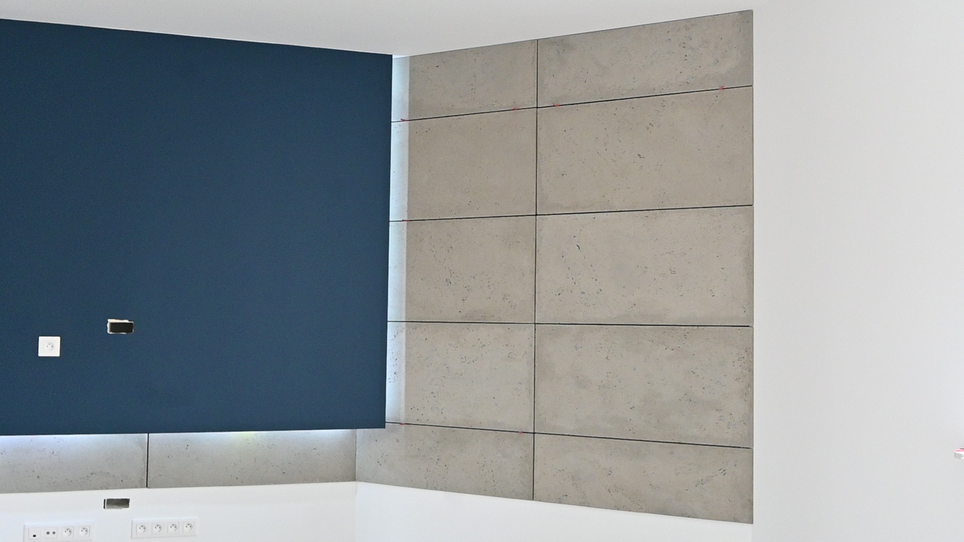 Narożnik pokoju po wykończeniu: ściana w kolorze granatowym z gniazdkami i szara ściana wyłożona płytami imitującymi beton. Widoczne listwy przypodłogowe i oświetlenie LED.